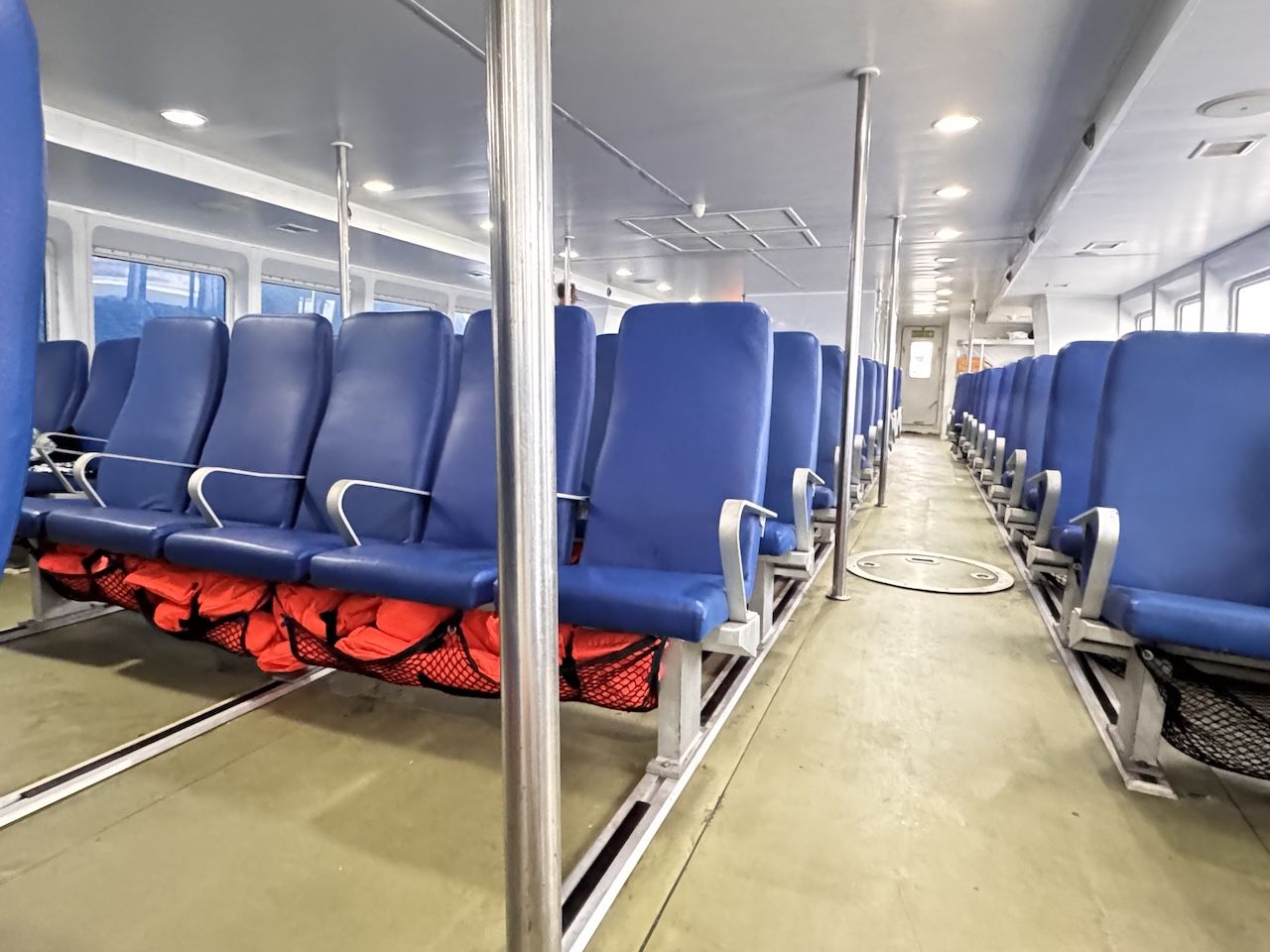 Leere Fährkabine mit blauen Sitzreihen, orangen Rettungswesten in Netzen und Mittelgang