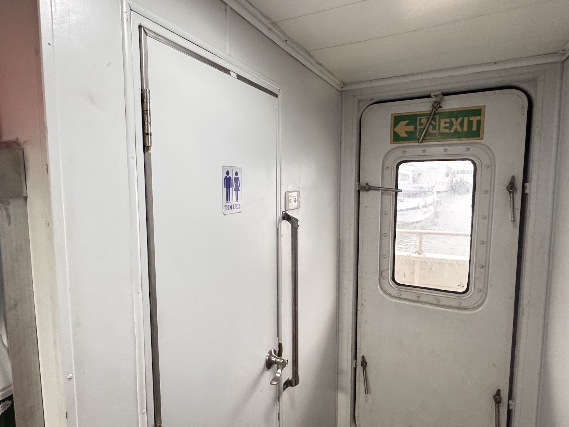 Gang einer Passagierfähre mit weißer Toilettentür und wasserdichter Ausgangstür mit grünem EXIT-Schild