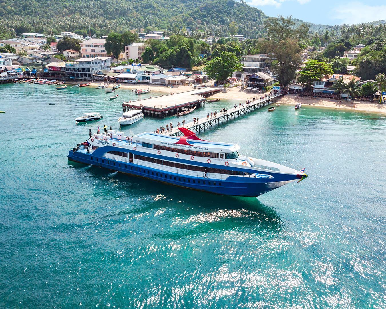 Aerial view of a blue and white ferry approaching a pier with tourists, tropical island bay
