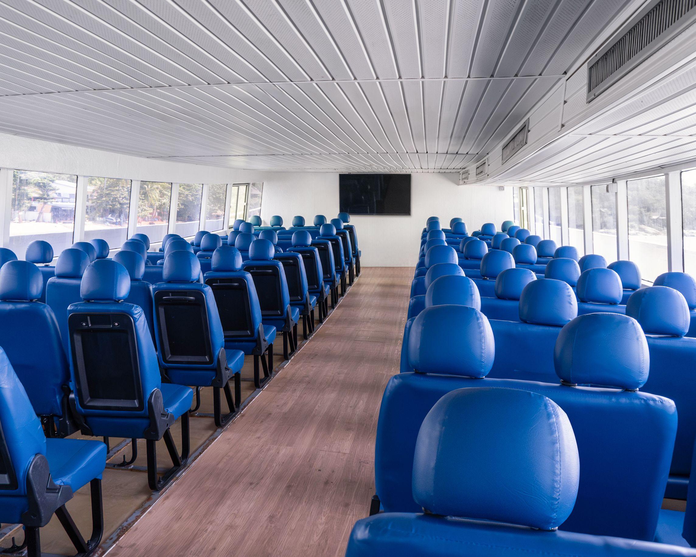 Empty passenger cabin of a ferry with rows of blue seats, wooden centre aisle and large windows