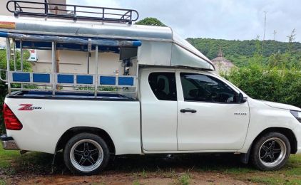 White pickup truck converted to a songthaew with bench seats, canopy and roof rack parked on grass by green hills
