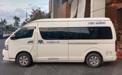 White minivan with Thai lettering on windows parked kerbside outside a building, palm trees behind