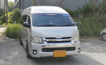 Silver Toyota Hiace high-roof minivan parked on a paved driveway beside grass and a modern building