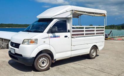 White Suzuki Carry pickup with covered passenger bench parked on a concrete pier beside calm tropical sea