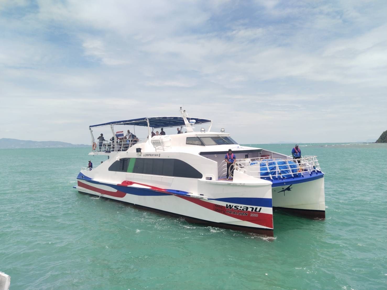 Lomprayah high-speed catamaran ferry with red, white and blue livery anchored on turquoise sea near a headland