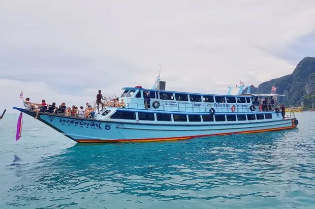 Blue and white Thai passenger ferry with sunbathers on the bow deck crossing turquoise sea near limestone cliffs