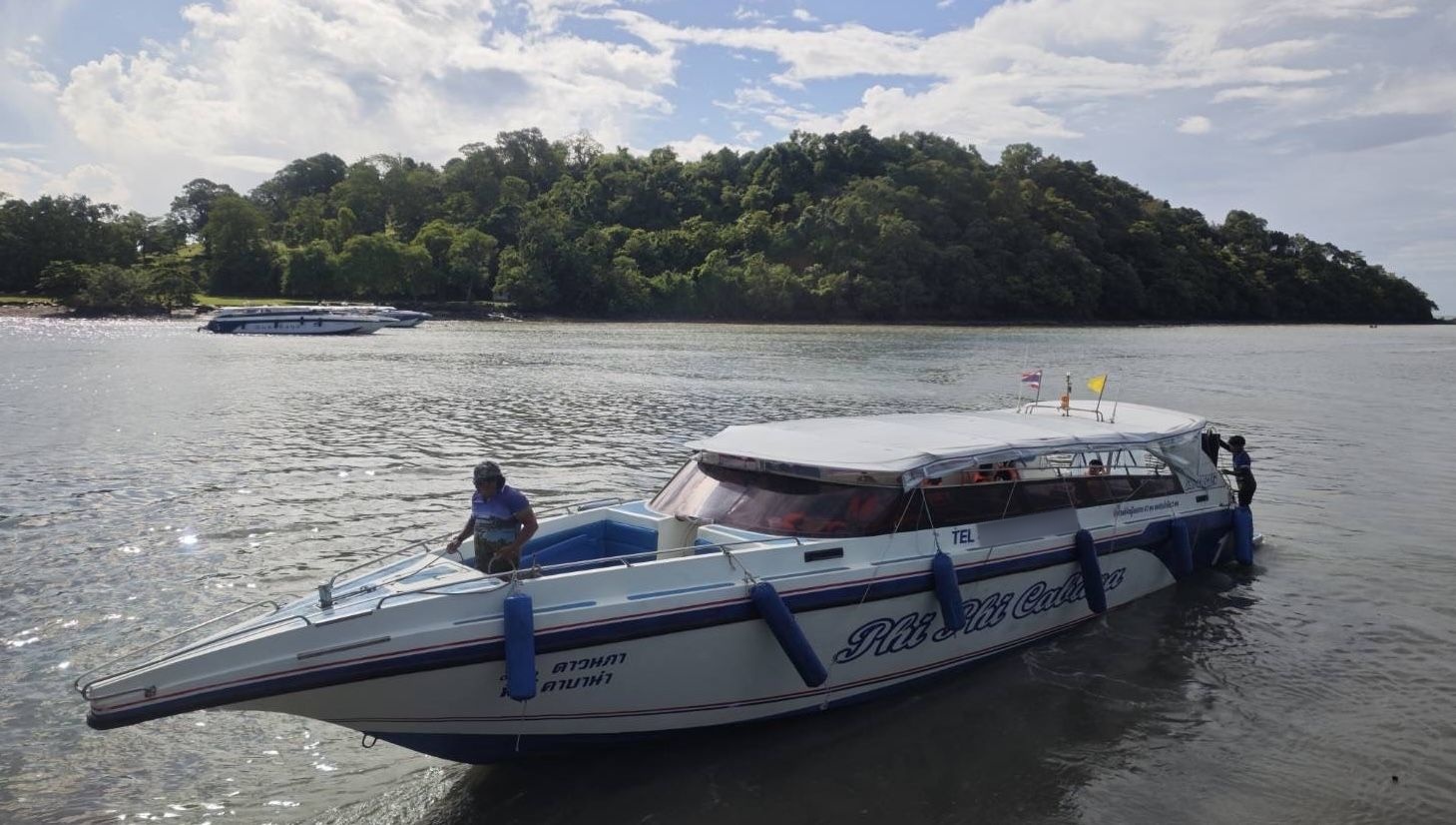 Weißes Schnellboot mit thailändischer Schrift im flachen Wasser nahe bewaldeter Insel, teils bewölkter Himmel