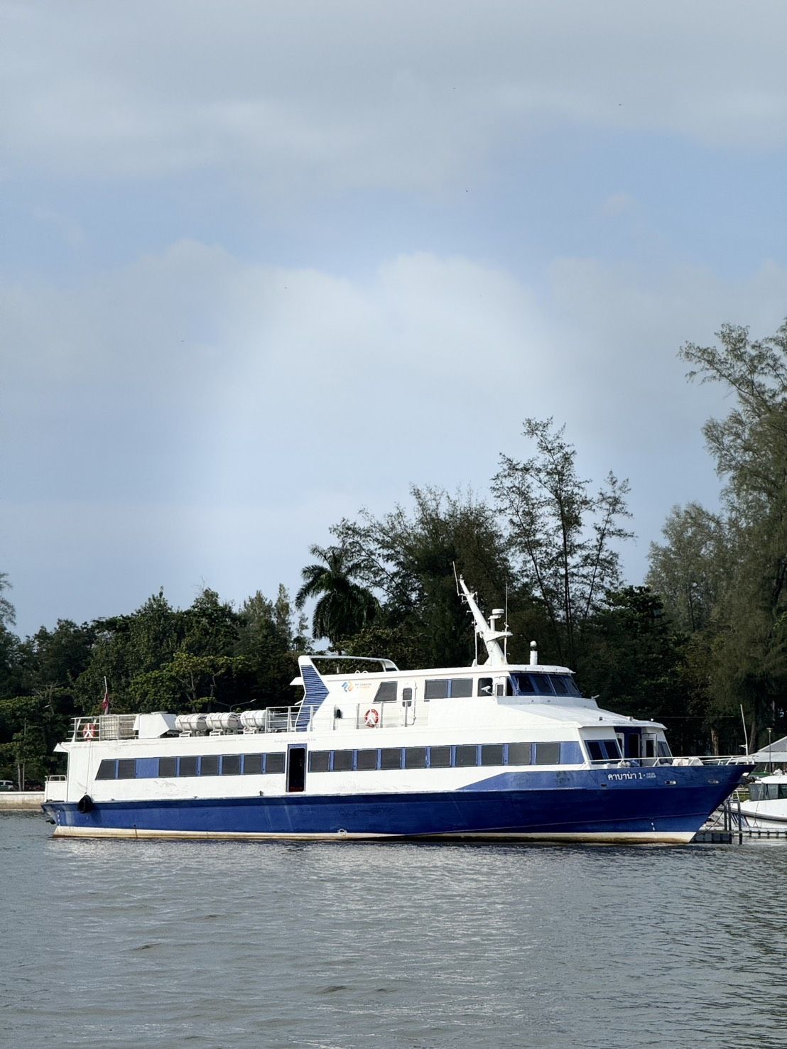 Blau-weiße Passagierfähre am Pier, thailändische Schrift am Bug, dahinter Kiefern