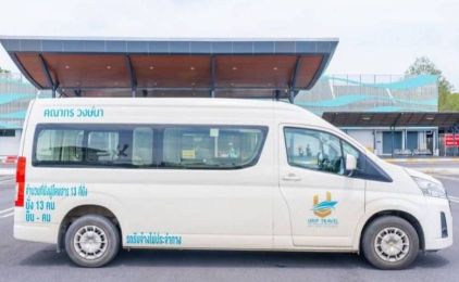 White high-roof minivan with Thai lettering and travel company logo parked under a terminal canopy