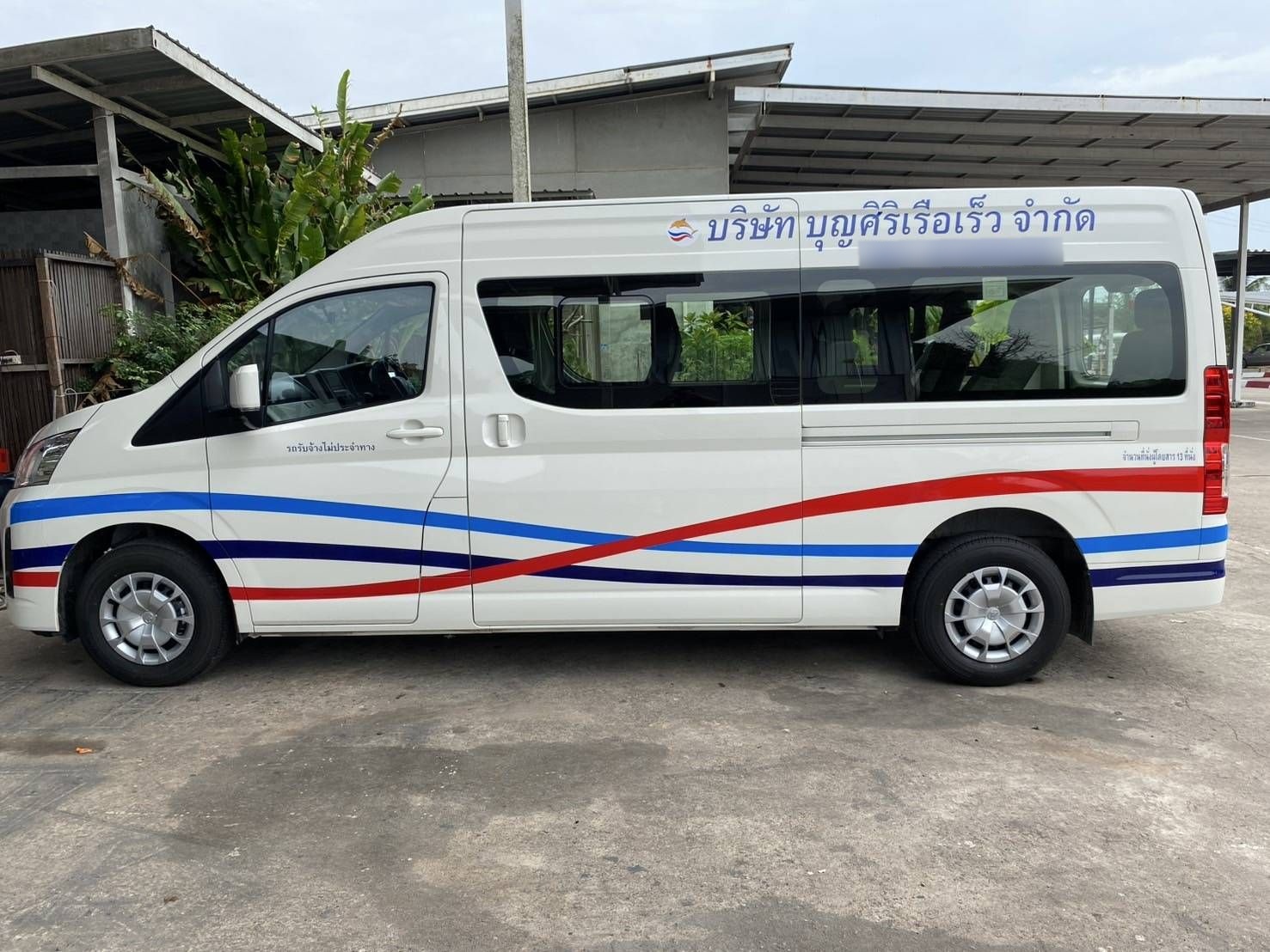 Weißer Minibus mit roten und blauen Schwungstreifen und thailändischem Firmenschriftzug auf Beton vor einem Carport