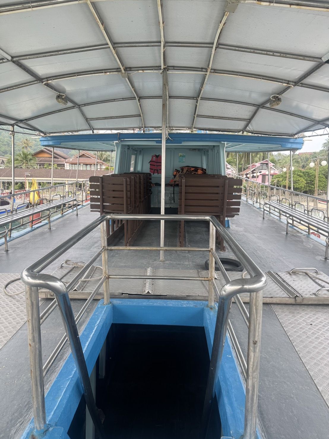 Leeres Oberdeck einer Fähre mit weißem Sonnendach, gestapelten Holzbänken und Treppe zum Unterdeck am Pier