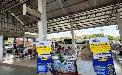 Open-air ferry terminal with blue check-in booths, covered waiting area, and boats visible in background