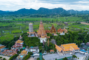 Aerial view of Wat Tham Sua temple complex with golden pagoda and tiered roofs beside green rice fields and hills