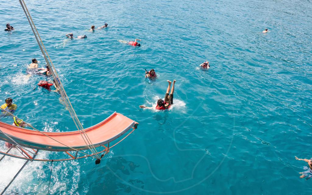 Snorkellers in red life jackets swim in turquoise sea beside an orange boat slide on a day-trip vessel