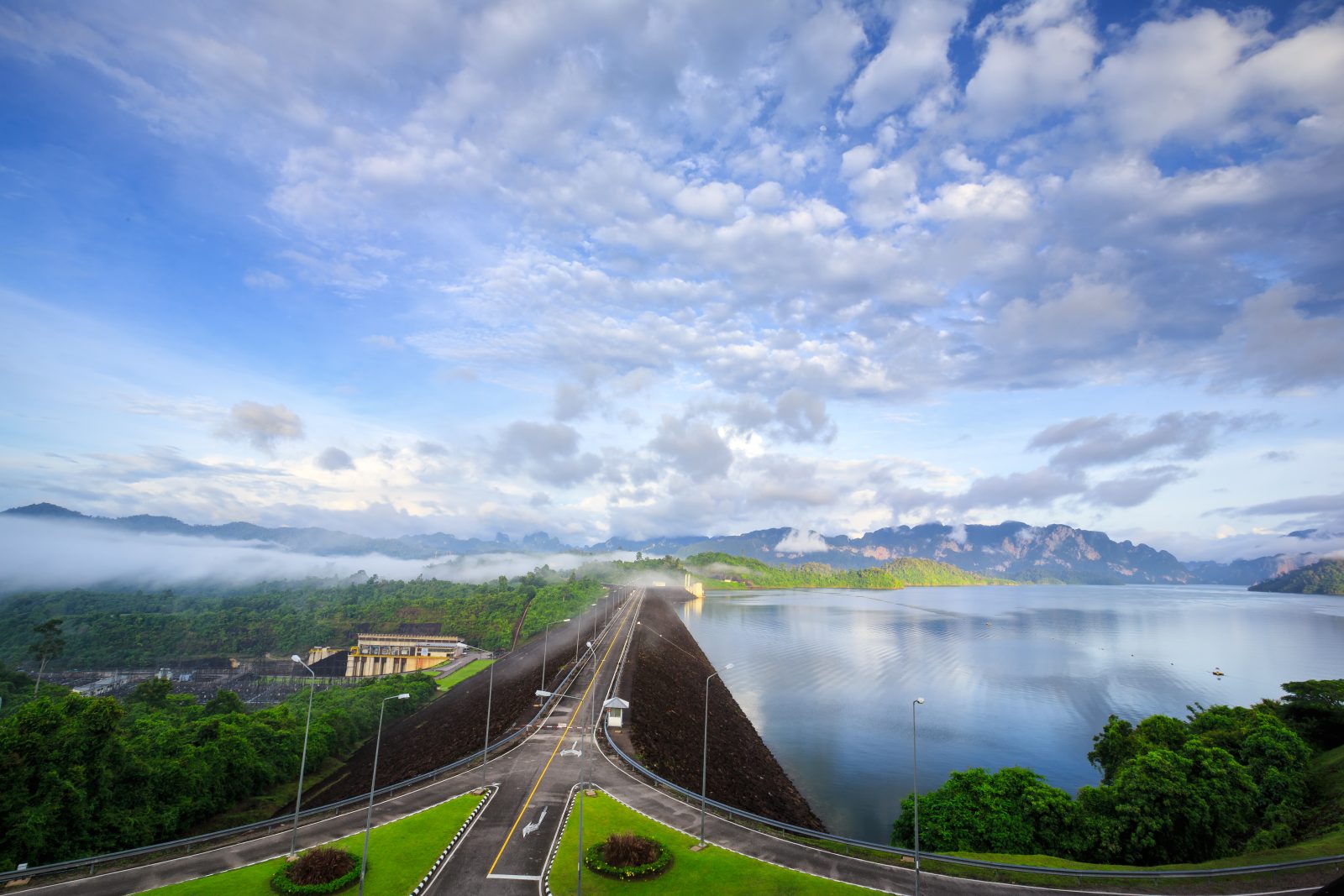 Road along a hydroelectric dam crest beside a calm reservoir with misty green mountains under blue sky